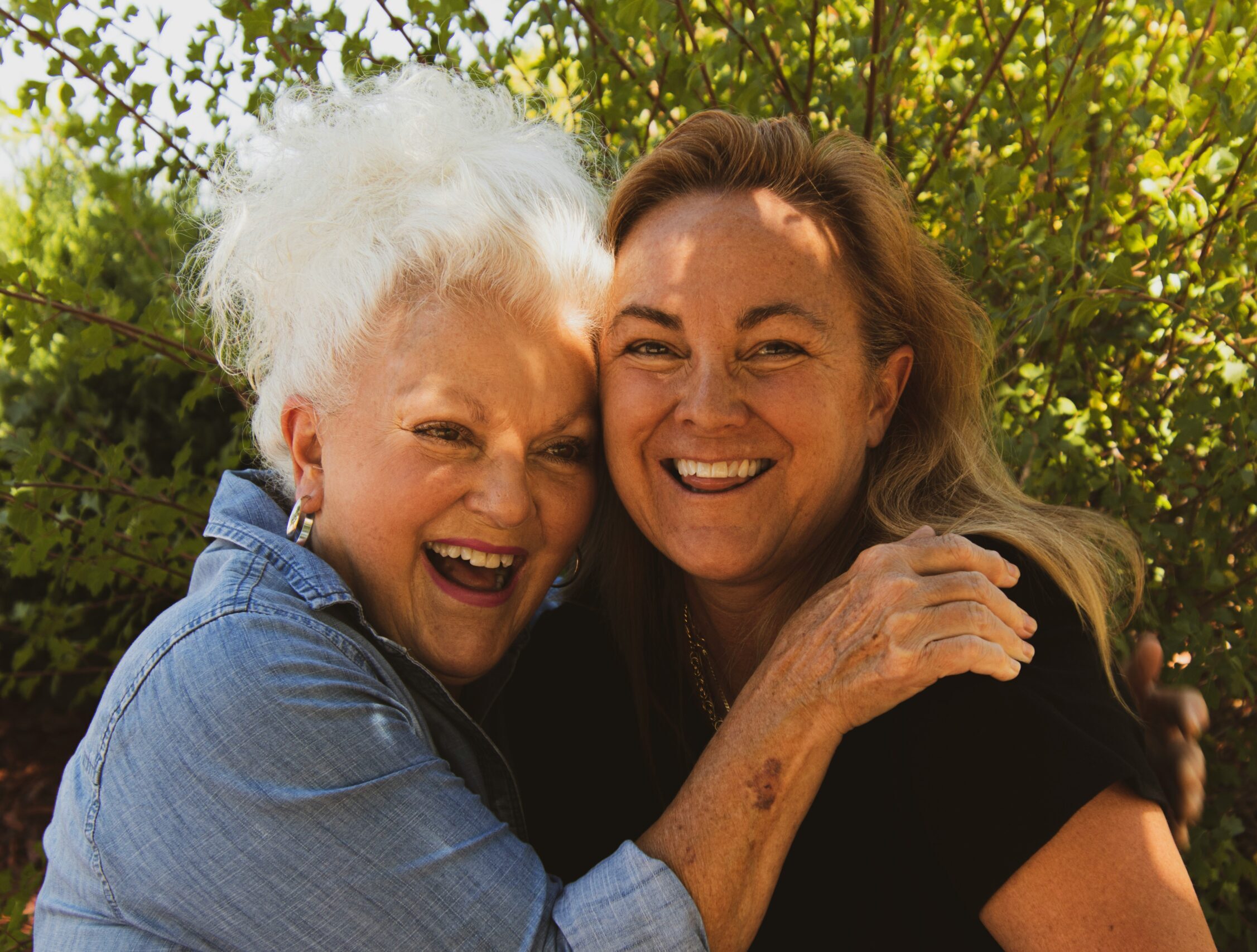 Starija žena i njegovateljica nasmijano se grle na otvorenom, prikazujući povjerenje i bliskost.