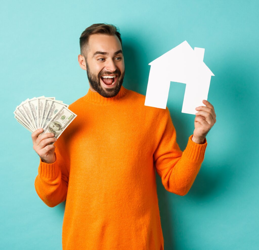 Našmijani muškarac u narančastom puloveru drži novčanice i papirnati model kuće, simbolizirajući usporedbu troškova.